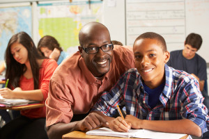 Teacher Helping Male Pupil Studying At Desk In Classroom